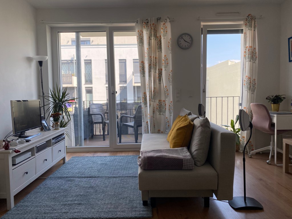An empty living room in an apartment in Berlin that shows a TV cabinet, a sofa, a study table and chair, and big glass windows with a balcony outside.